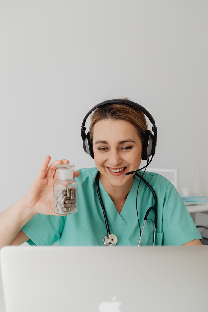 about-02 Nurse in green scrubs wearing headphones smiles while holding a pill bottle during a video call.